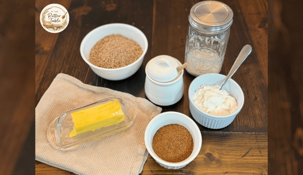 Six staple kitchen ingredients including butter, wheat berries, sourdough starter, Greek yogurt, honey, and coconut sugar arranged on a wooden table.